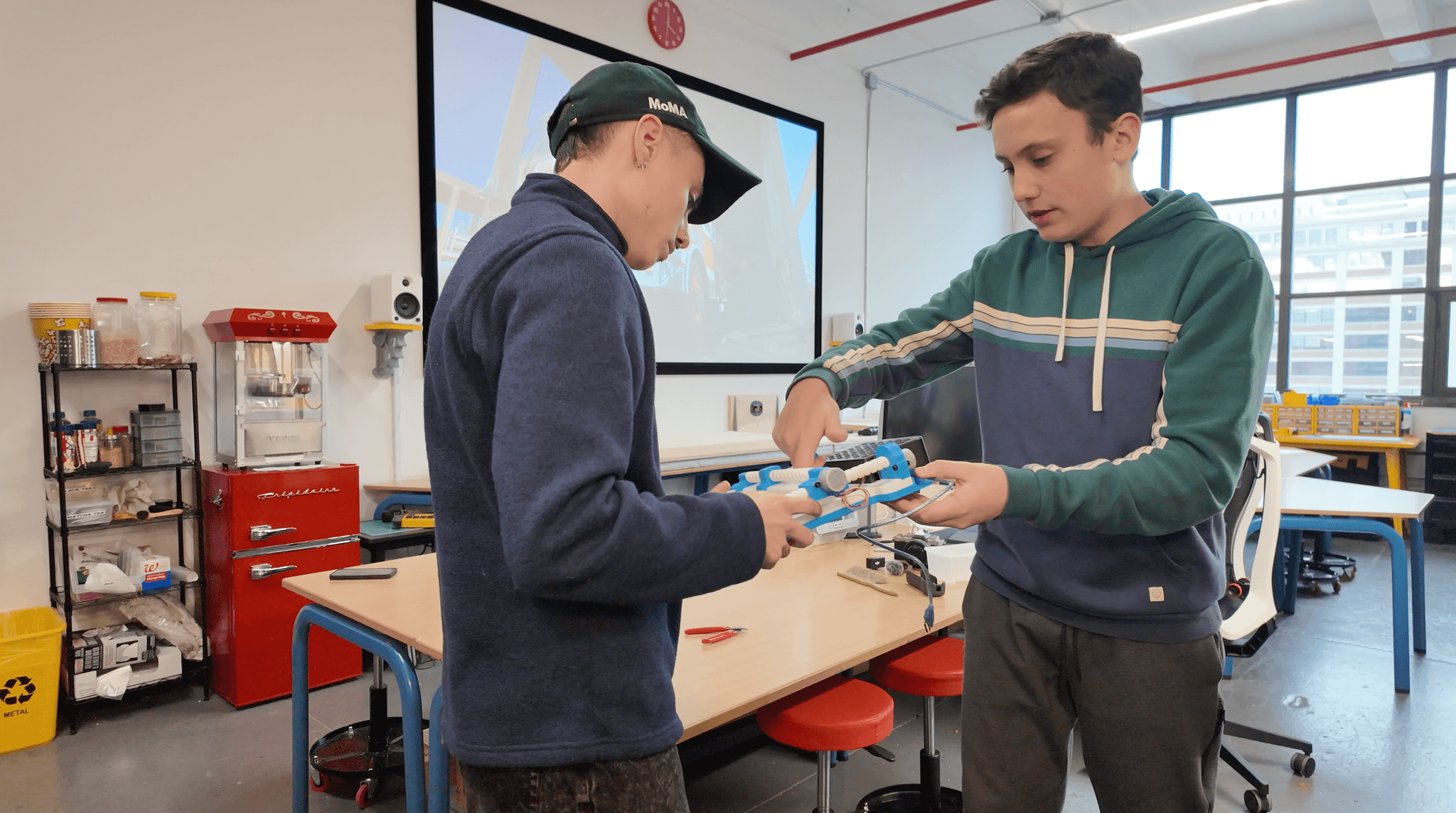 A mentor explaining a hand-built mechanical prototype to a young maker at Desk Job Design Lab